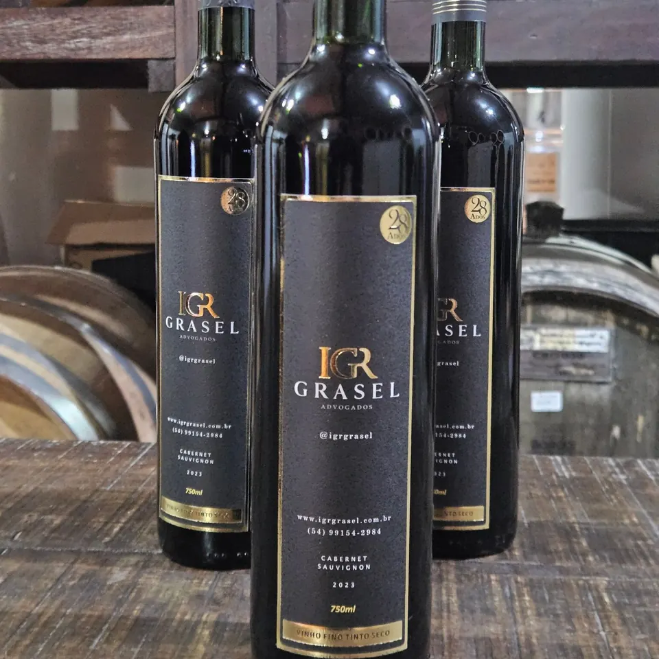 Três garrafas de Cabernet Sauvignon com rótulo preto e dourado personalizado de um escritório de advocacia, na cave entre barris de carvalho